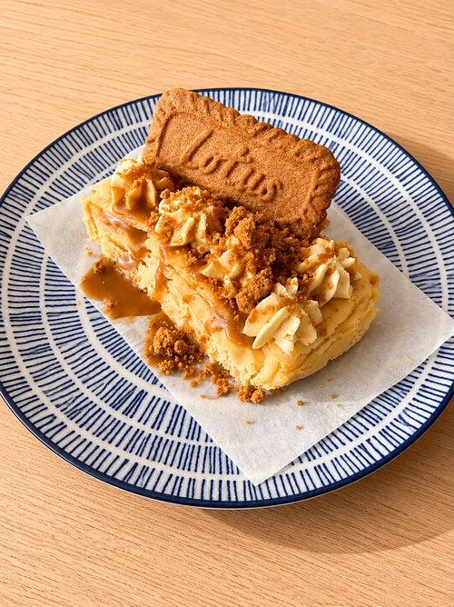 A slice of Lotus caramel and biscuit cheesecake on a blue and white patterned plate, set on a light wooden table.