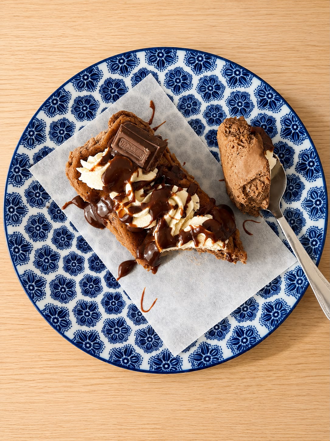 A slice of chocolate cheesecake with whipped cream, chocolate drizzle, and a chocolate bar, served on parchment paper with a spoonful of chocolate on a blue and white patterned plate over a light wood table.