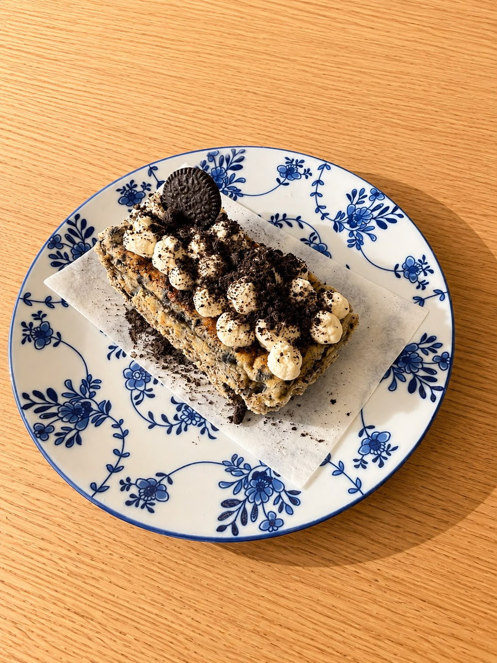 A slice of basque cheesecake topped with cookie crumbles and white chocolate drizzle, served on a white ceramic plate with a blue floral pattern over a sheet of parchment paper, set on a wooden table.