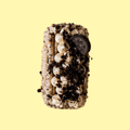 An Oreo-themed basque cheesecake with white frosting, dark chocolate cookie crumbs, and a whole Oreo cookie on top, presented on a solid pale yellow background.