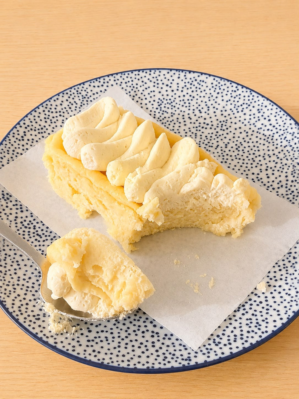 A close-up of a slice of creamy cheesecake with piped cream on a speckled blue and white plate, with a spoonful of frosting in front on a light wood surface.