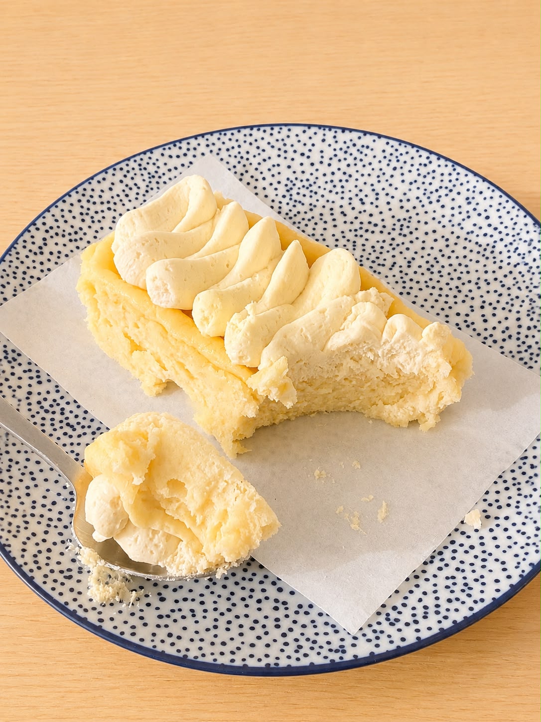 A close-up of a slice of creamy cheesecake with piped cream on a speckled blue and white plate, with a spoonful of frosting in front on a light wood surface.